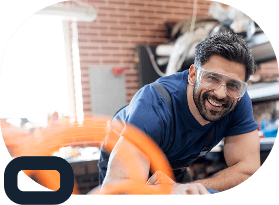 Smiling man wearing safety goggles and a blue shirt working in a workshop with brick walls, leaning over a workbench with blurred orange tool in foreground.
