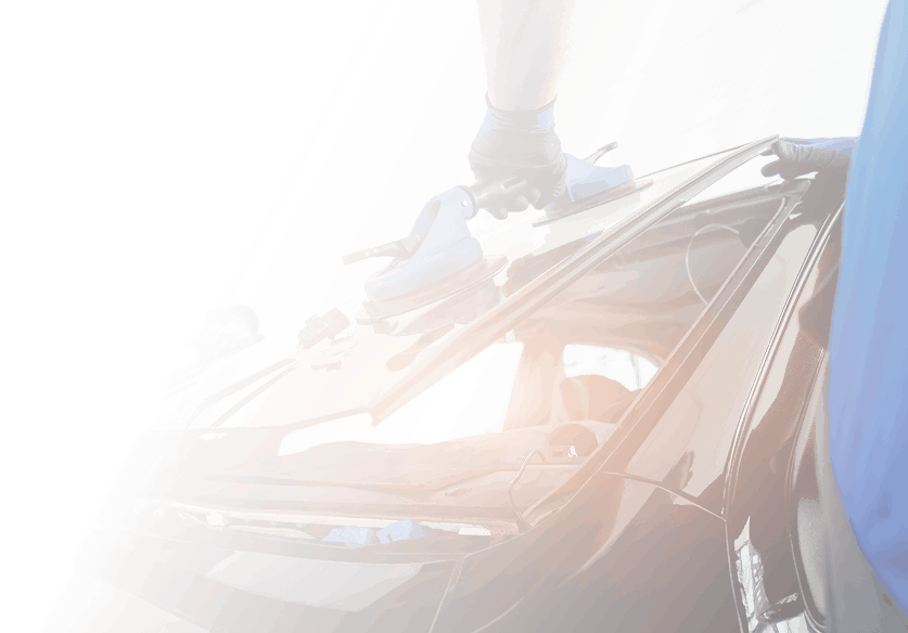 A worker installing or repairing a car windshield, using suction cups to handle the glass on a black vehicle.